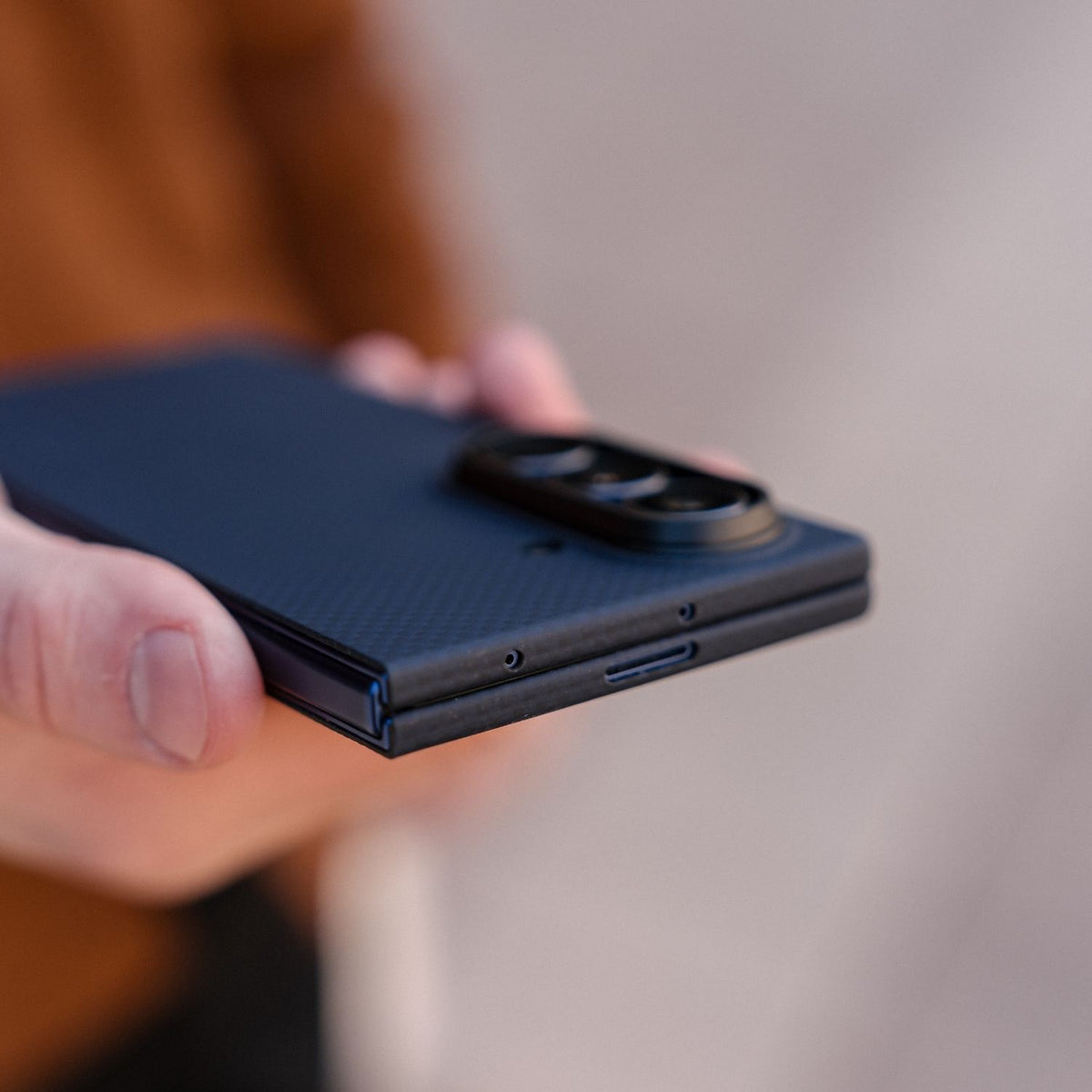 A close-up of a person holding a blue carbon-fiber-textured galaxy z fold 8 ultra case, showing the outer back cover and part of the hinge.