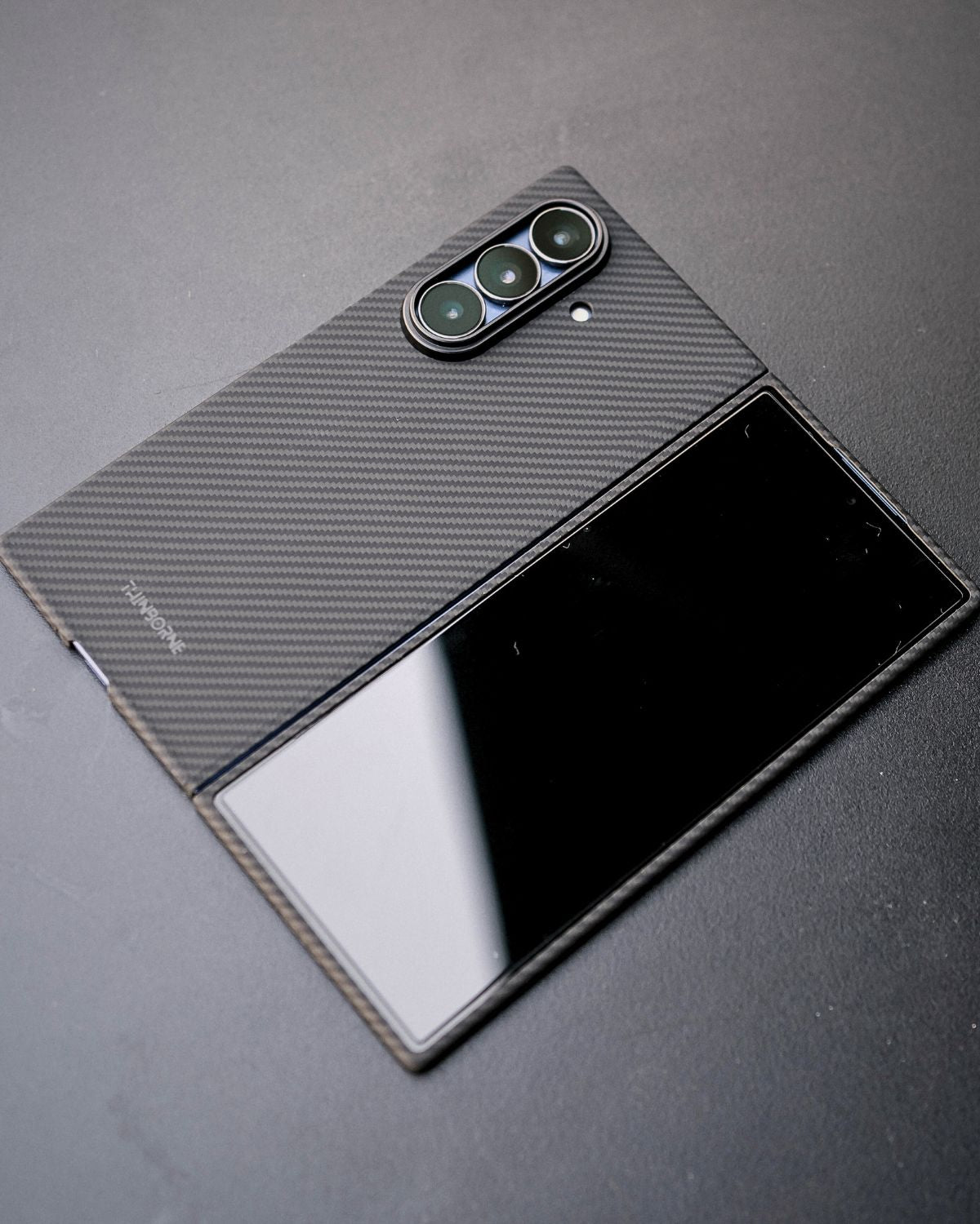  A close-up of a closed, black, foldable phone. The back of the phone is covered with a black carbon fiber case. The triple camera lenses are prominent on the left side of the back.


