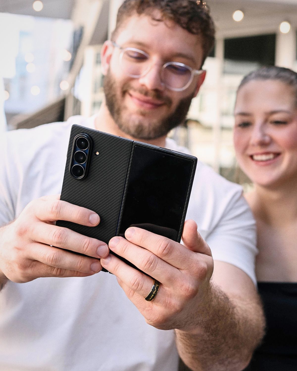 A person holds a black foldable phone with a carbon fiber case. The phone is unfolded and held in both hands. The back of the phone shows the triple camera setup.

