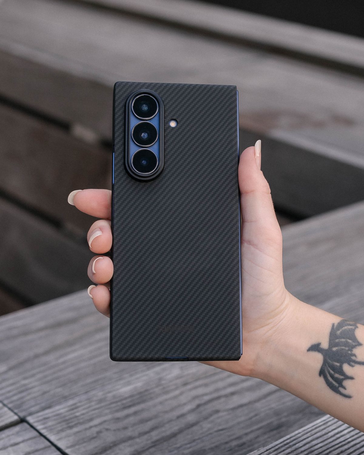 A person's hand holds a black foldable phone with a carbon fiber case. The phone is closed, and the camera lenses are clearly visible on the back. The carbon fiber pattern is visible.