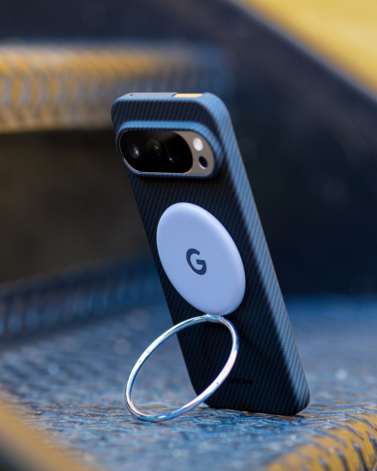 A phone with a dark blue, textured Pixel 10 Pro xl case stands on a metal grate, using a gray magnetic ring accessory as a kickstand.