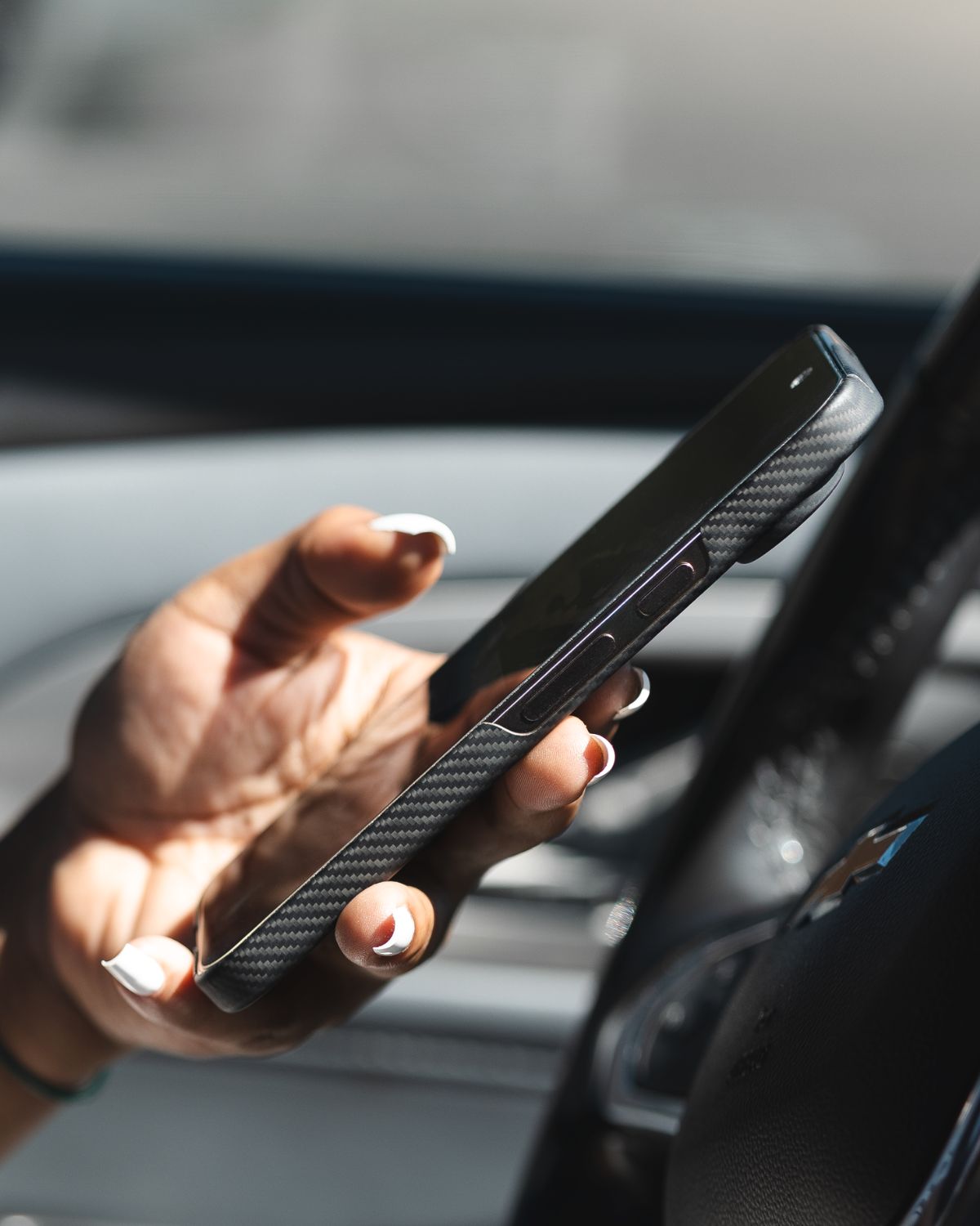 A person's hand with white manicured nails holds a phone in a black, textured Pixel 10 Pro xl case inside a car, with the steering wheel visible in the background.