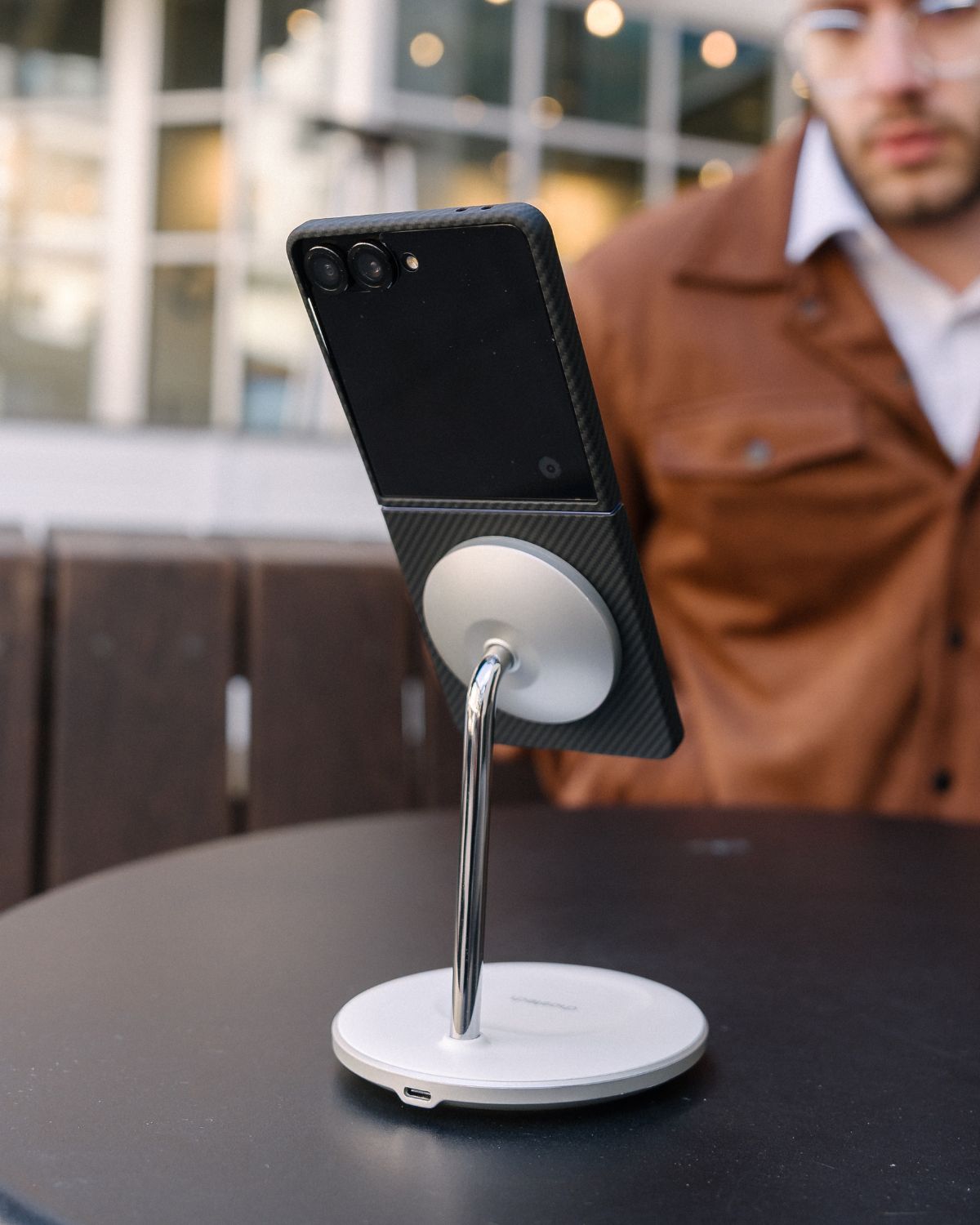 galaxy z flip 7 with magssafe case attached to a Phone holder on a table with a blurred background of a person and building