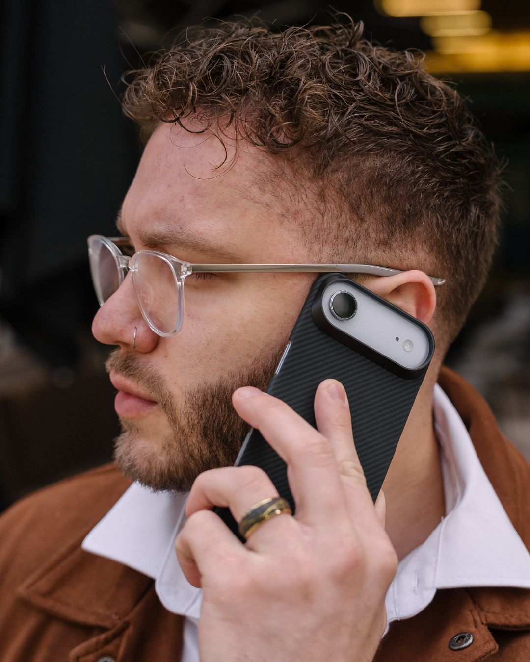 Man with glasses holding a phone with iPhone air case to his ear, wearing a brown jacket.