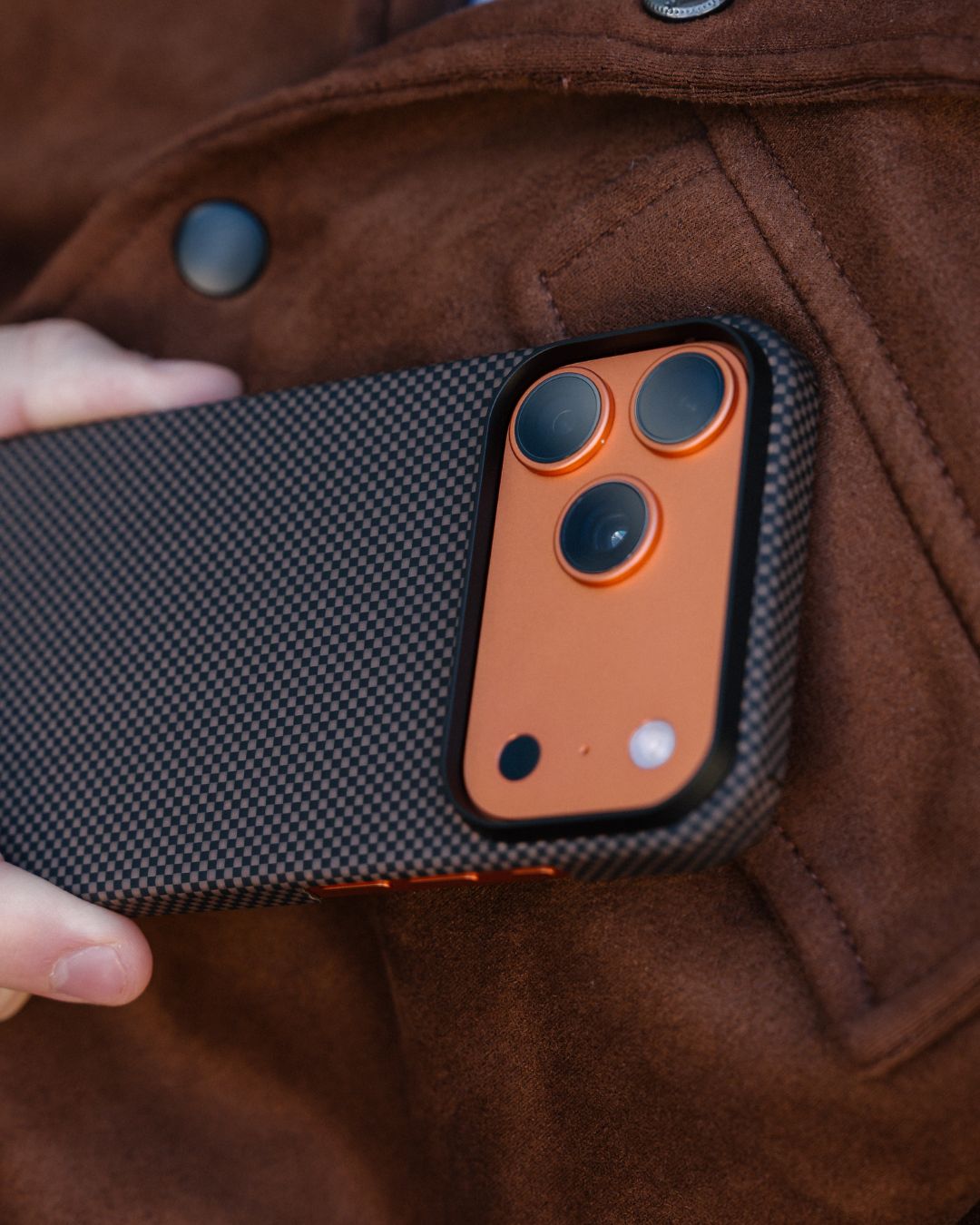 iphone 17 pro max case with carbon fiber texture and orange accents held against a brown jacket.