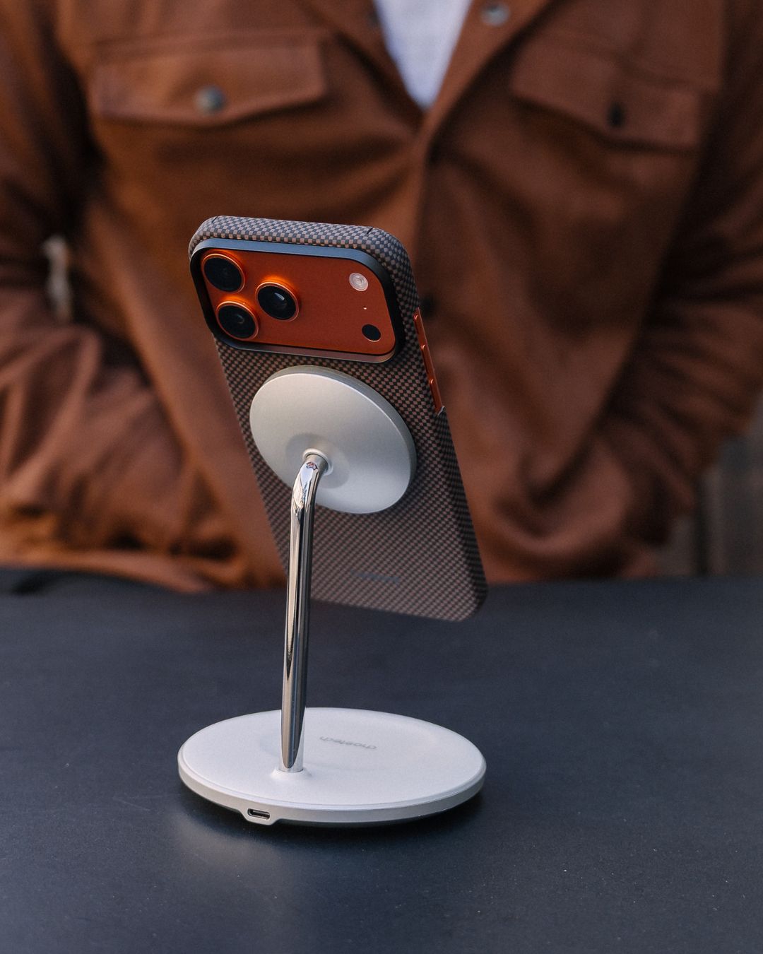 Magsafe stand with a iPhone 17 pro max with a phone on top, placed on a dark surface with a blurred brown jacket in the background.