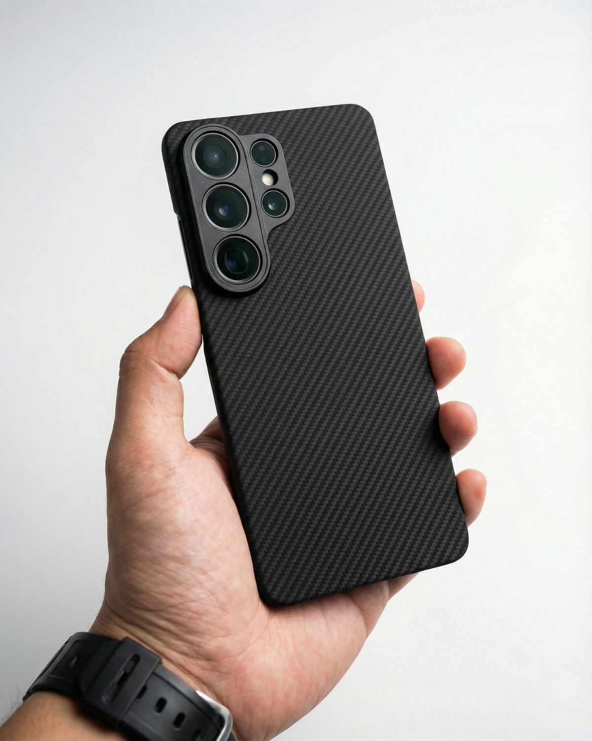 Hand holding a black samsung galaxy s26 ultra phone case with a carbon fiber texture on a light gray background