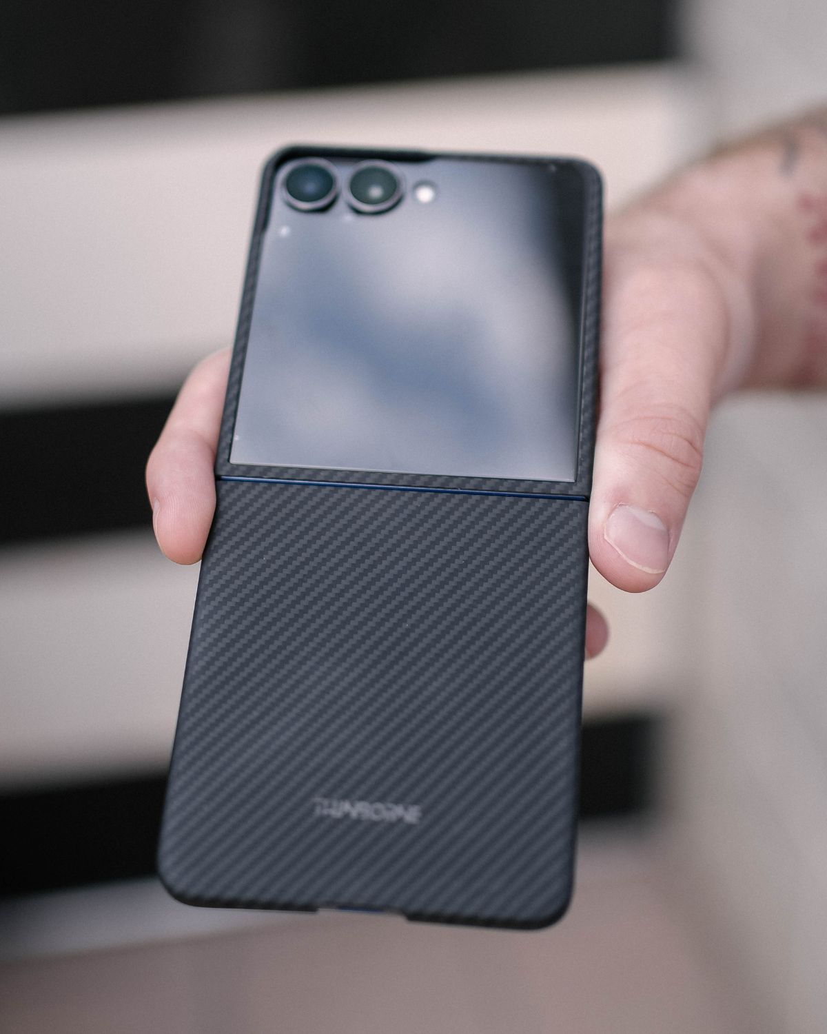 A hand holds a partially folded Samsung Galaxy Z Flip 7 with a thin carbon fiber case, showing the 