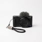 Sony ZV-E T3 camera in black with a gray furry microphone windscreen and a black wrist strap with a silver metal connector, photographed on a clean white background.