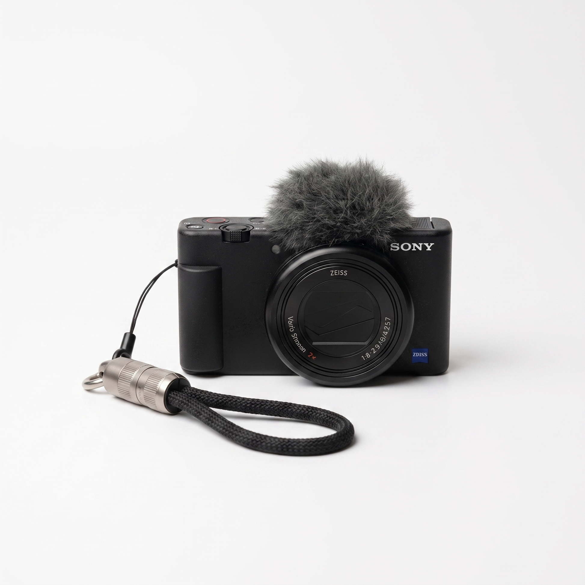 Sony ZV-E T3 camera in black with a gray furry microphone windscreen and a black wrist strap with a silver metal connector, photographed on a clean white background.