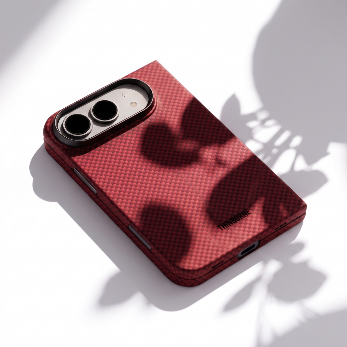 A red iphone duo phone case with a black camera bezel, shown at an angle on a white surface with artistic leaf shadows.