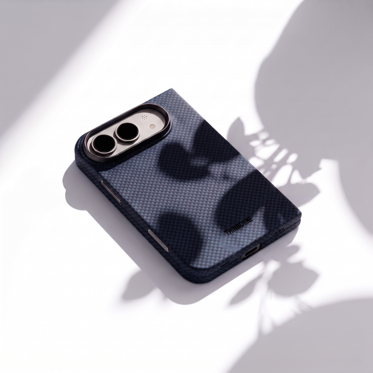 A dark navy blue iphone duo phone case with a textured carbon-fiber-like pattern and a black camera bumper, shown on a white surface with artistic leaf shadows.