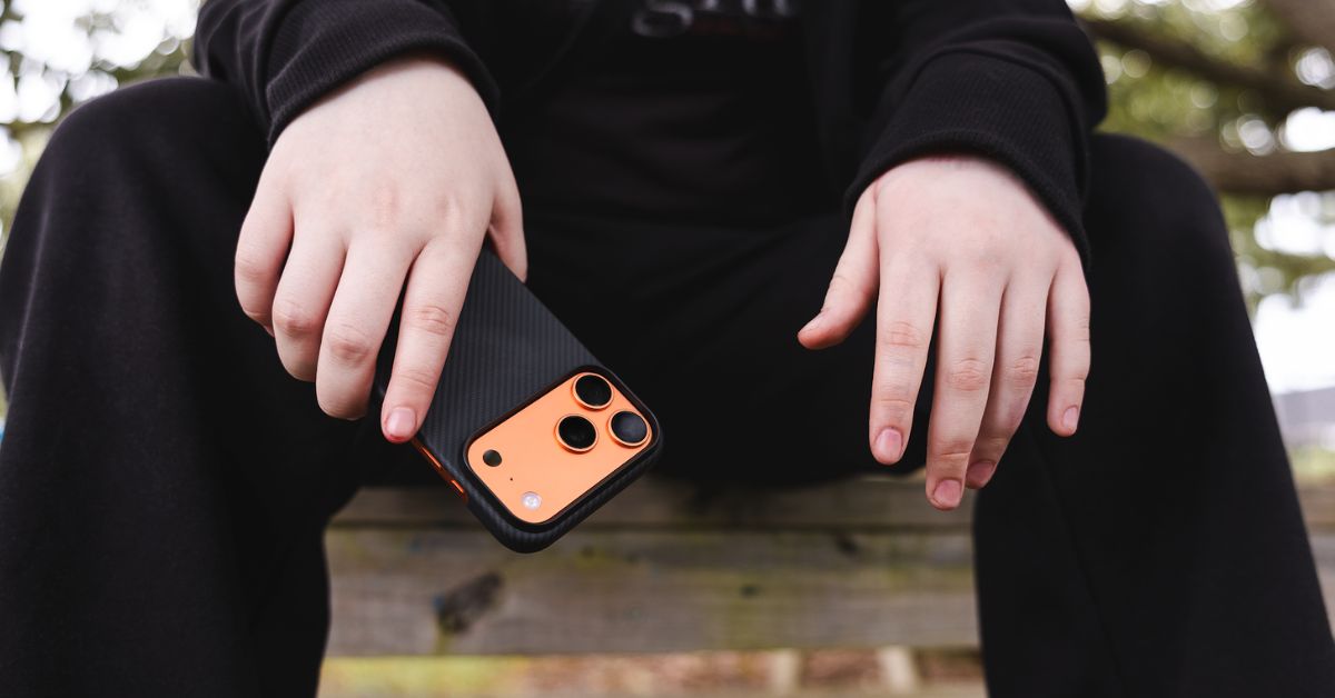 Person holding an iPhone 17 Pro Max with a slim black aramid fiber case, showing the orange back and triple-camera cutout.