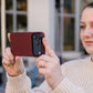Woman taking a photo in landscape mode with an iPhone 17 Pro Max in a rugged deep red textured case.