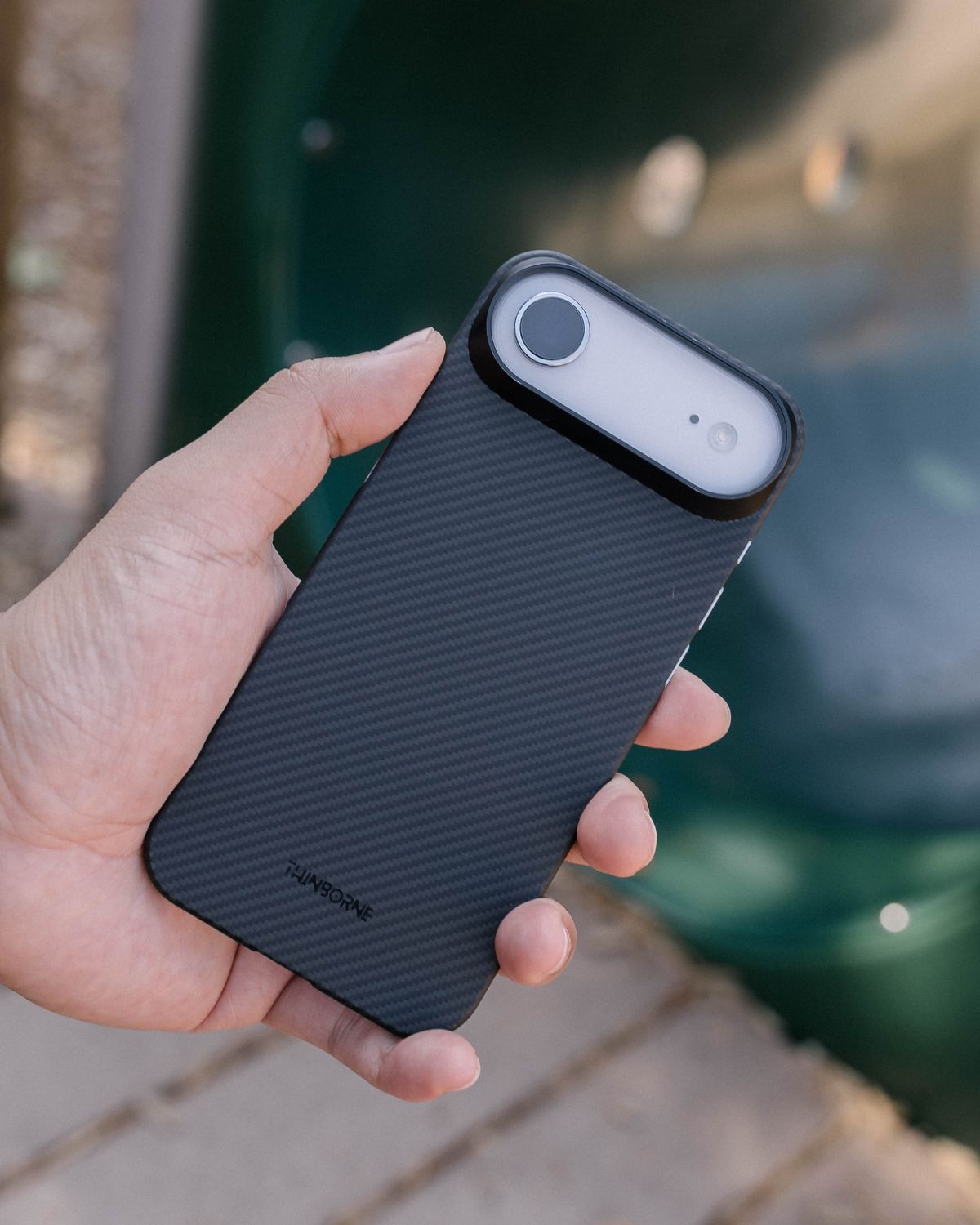 A person holds an iPhone with a black, carbon-fiber textured case. The large, pill-shaped camera cutout is visible at the top. 