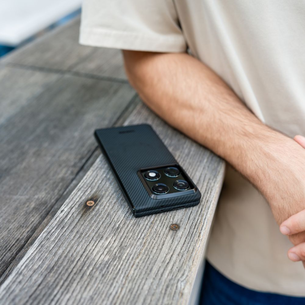 A close-up shot of a motorola razr fold in a black carbon fiber case, resting on a weathered wooden surface next to a person's arm.