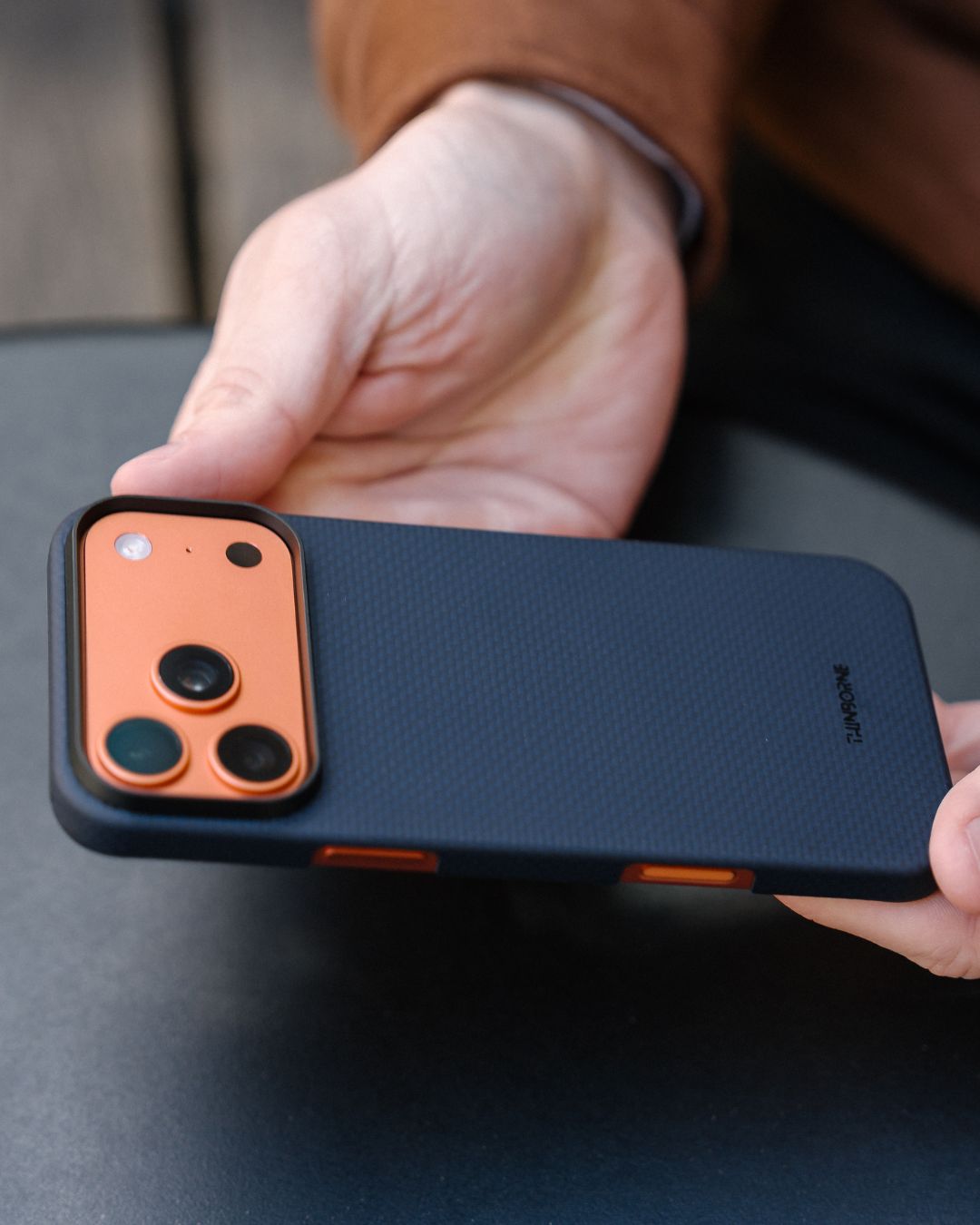 Person holding a iPhone 17 pro max with a navy case on a dark surface