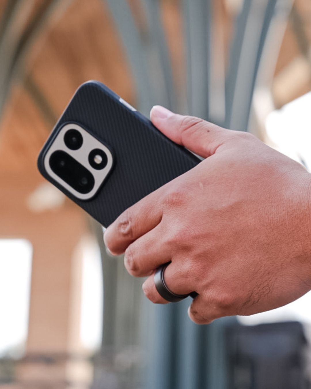A person holding the OnePlus 15 in the aramid fiber case outdoors. The light catches the textured weave of the material, and the silver accents of the camera module stand out against the matte black finish.
