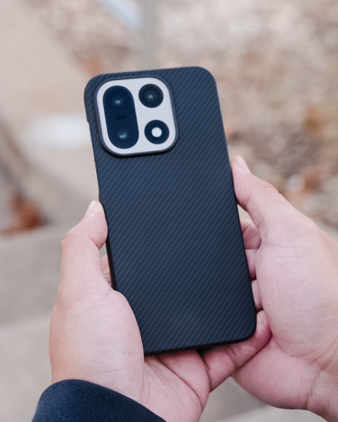 A person holding the OnePlus 15 in a slim, black aramid fiber case with a distinct carbon fiber-style diagonal weave. The case features a precision-molded silver-rimmed cutout for the phone's unique square-shaped camera module.
