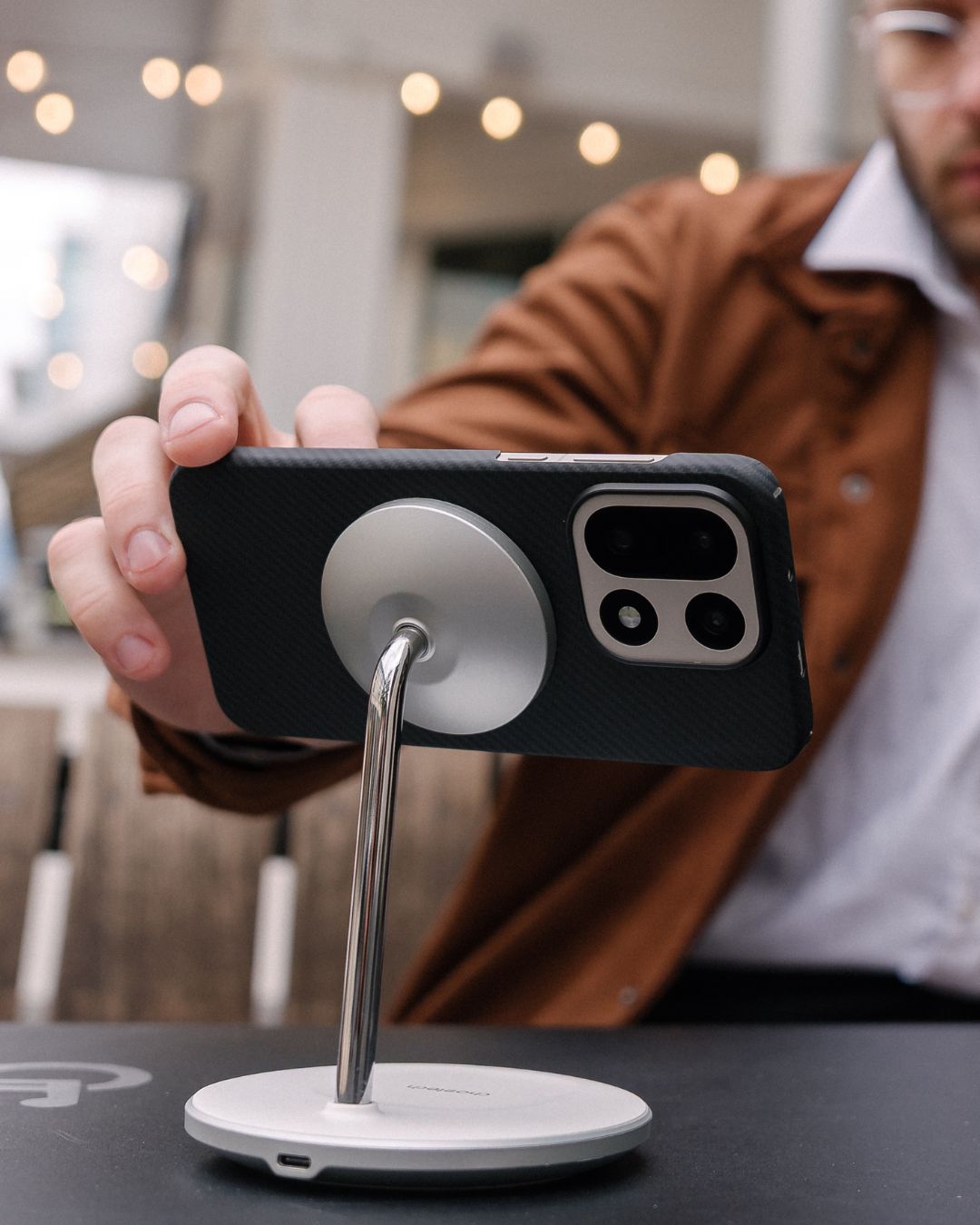 Black carbon fiber OnePlus 15 case with MagSafe compatibility attached to a silver magnetic charging stand in a landscape orientation.
