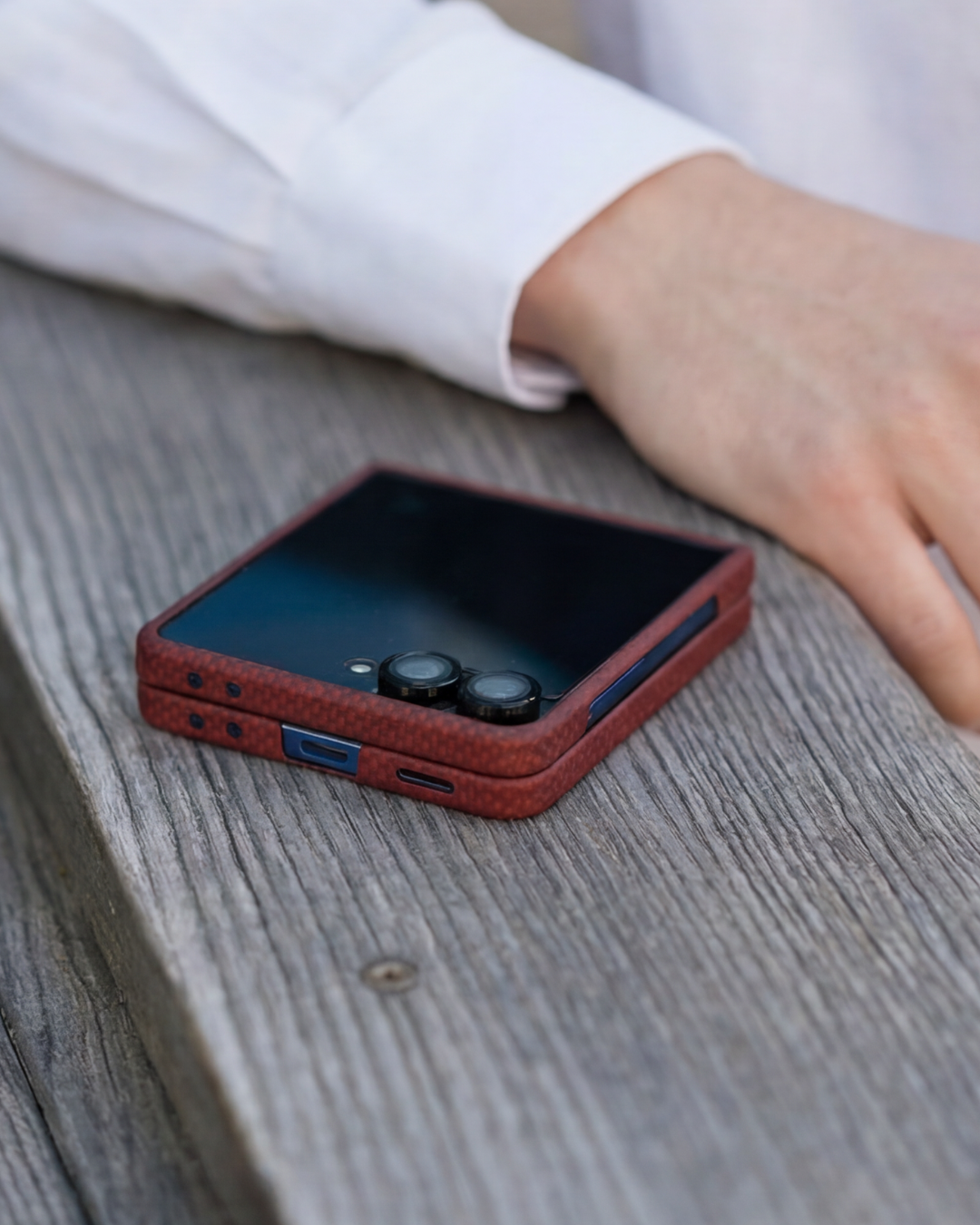 Phone with a red z flip 7 case on a wooden surface, with a person's hand partially visible.
