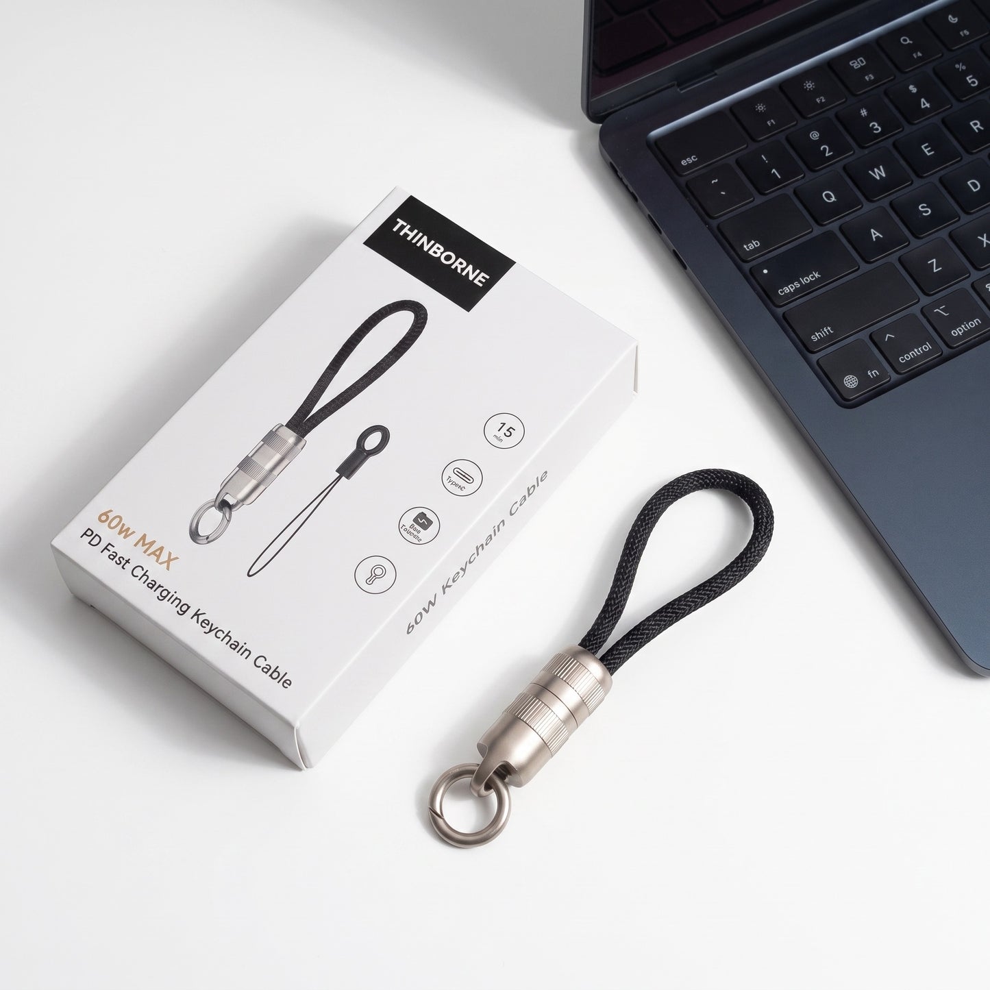 A mini PS/1 fast charging function cable with a black loop and silver metal connector, shown next to its white product packaging box and a dark laptop keyboard on a white surface.