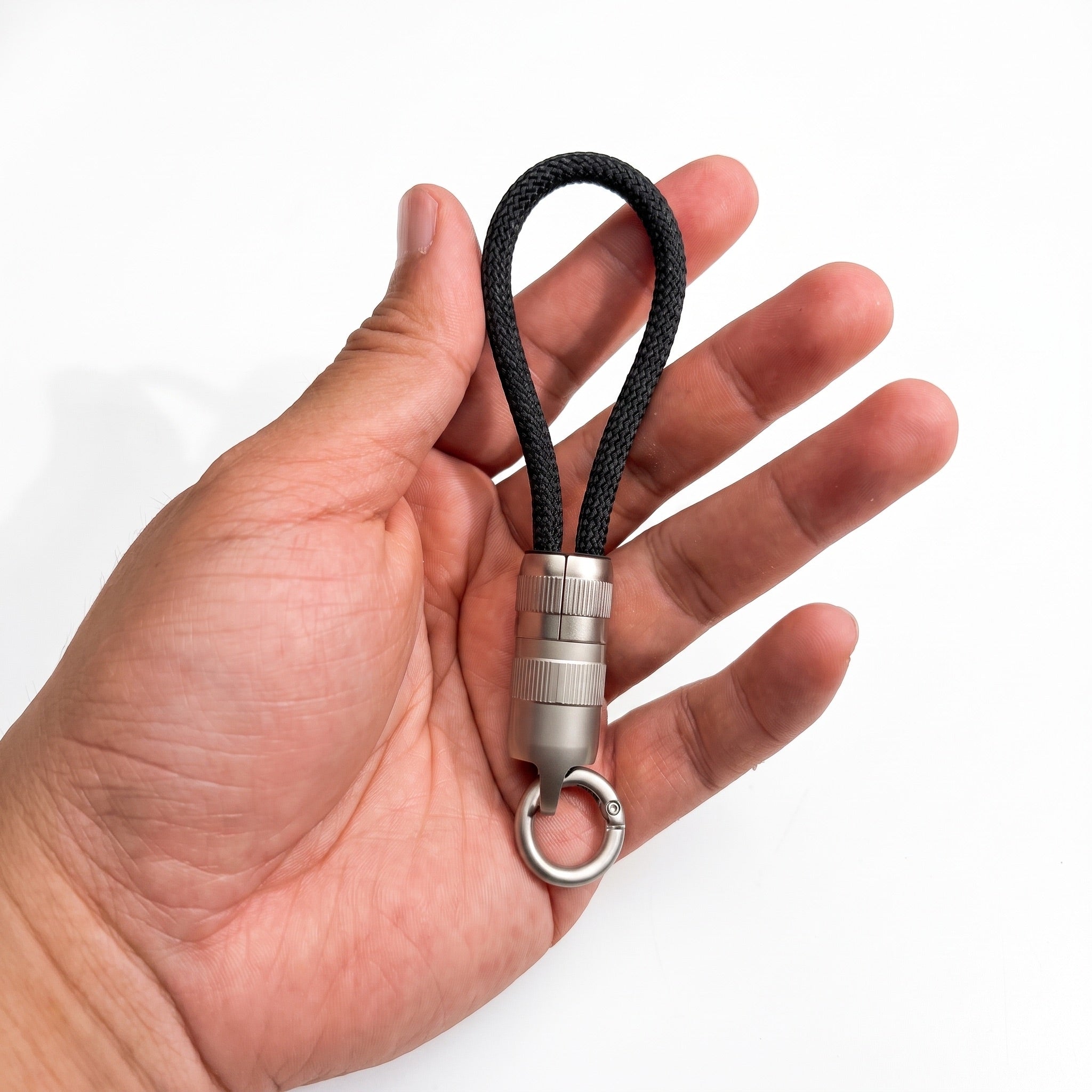 A hand holding a black braided cord keychain with a silver metal cylindrical connector and key ring, shown against a plain white background.