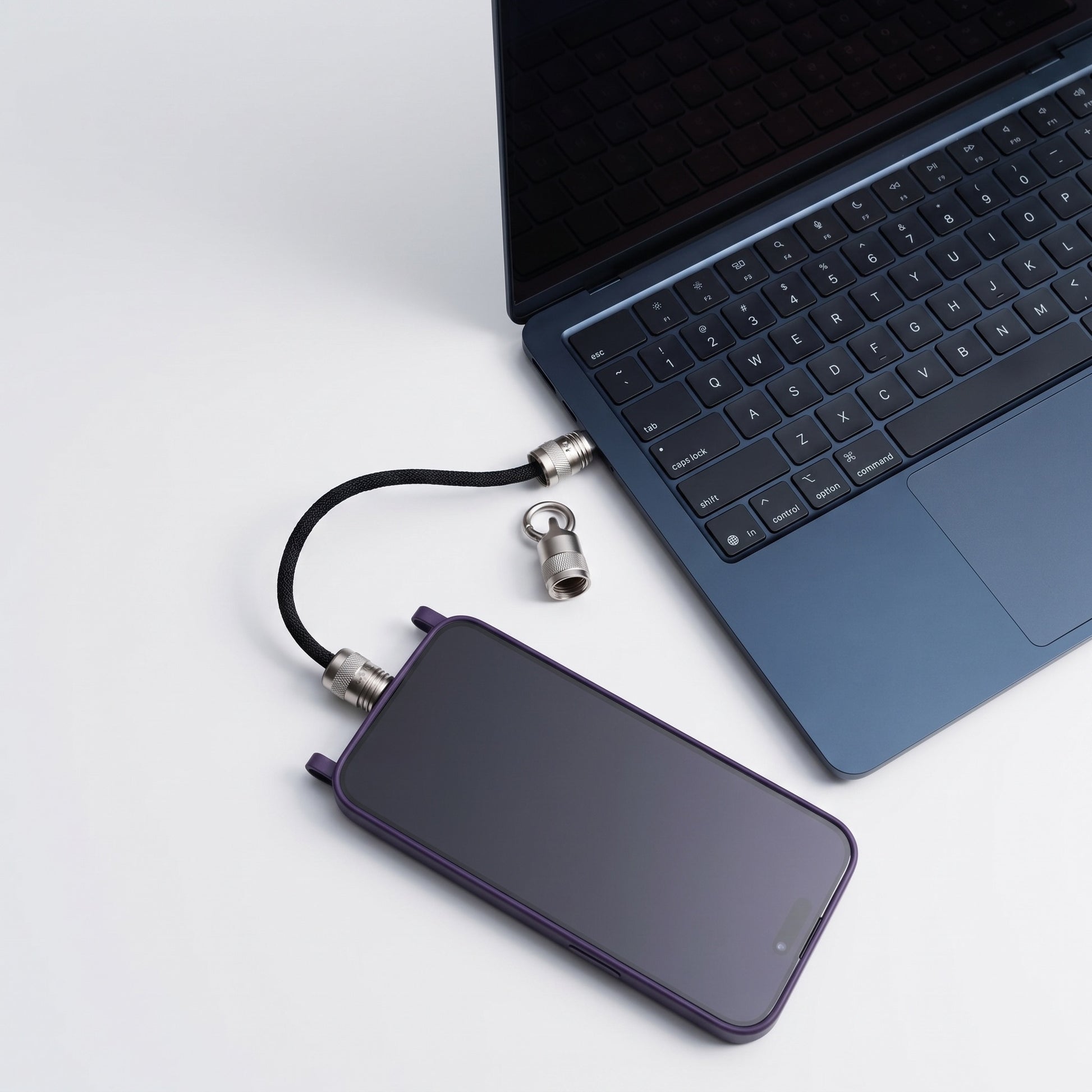 A purple smartphone with a black cable and silver lock connector lies next to an open blue laptop on a clean white surface.