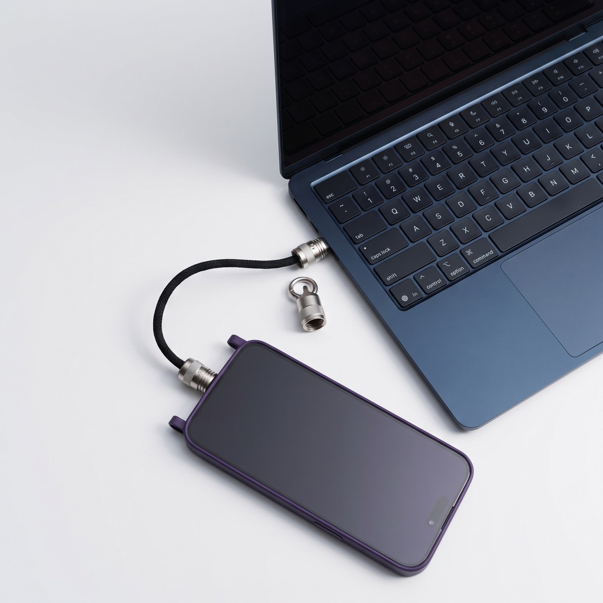 A purple smartphone with a black cable and silver lock connector lies next to an open blue laptop on a clean white surface.